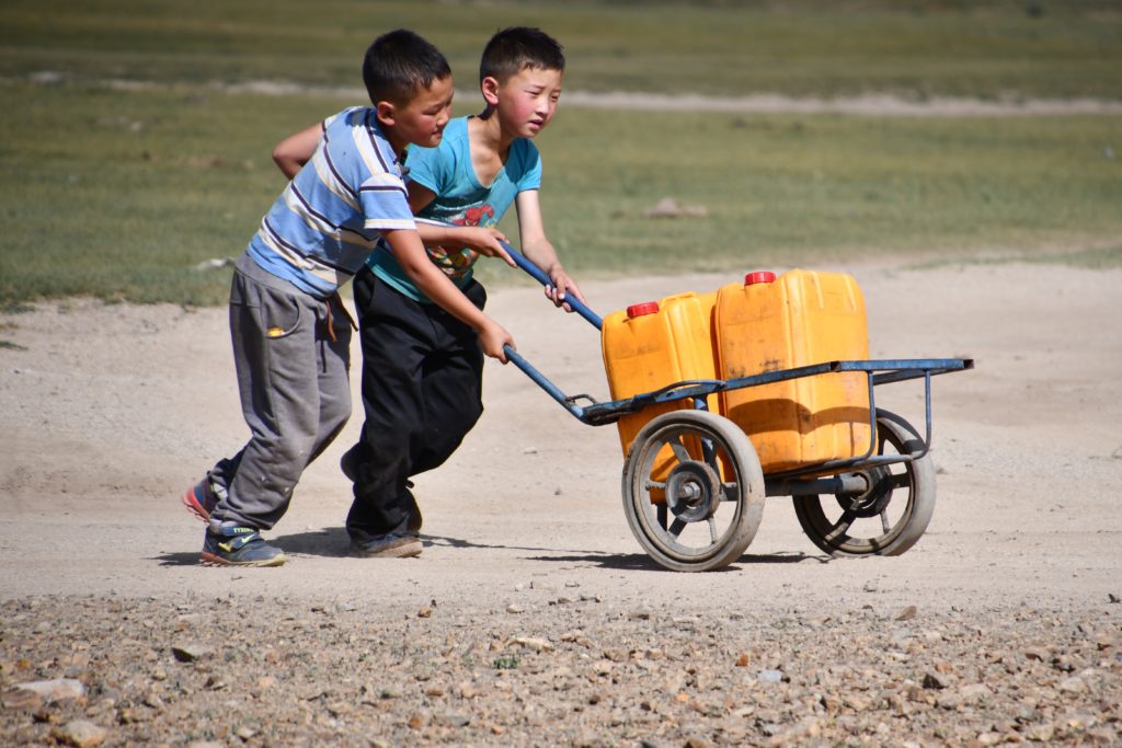 Two boys transporting two yellow water canisters on a wheelbarrow